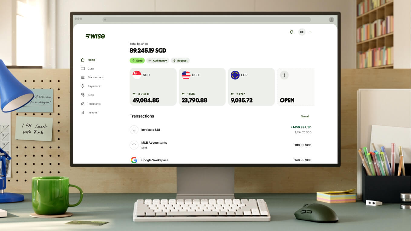 A close-up shot of a modern workspace shows a white computer monitor displaying the Wise dashboard. The screen shows a total balance of "89,245.19 SGD" and three main currency accounts: SGD with "49,084.85," USD with "23,790.88," and EUR with "9,035.72," followed by an option to open a new account. Recent transactions are listed below. In front of the monitor is a compact white keyboard, a dark grey mouse, and a bright green ceramic mug.